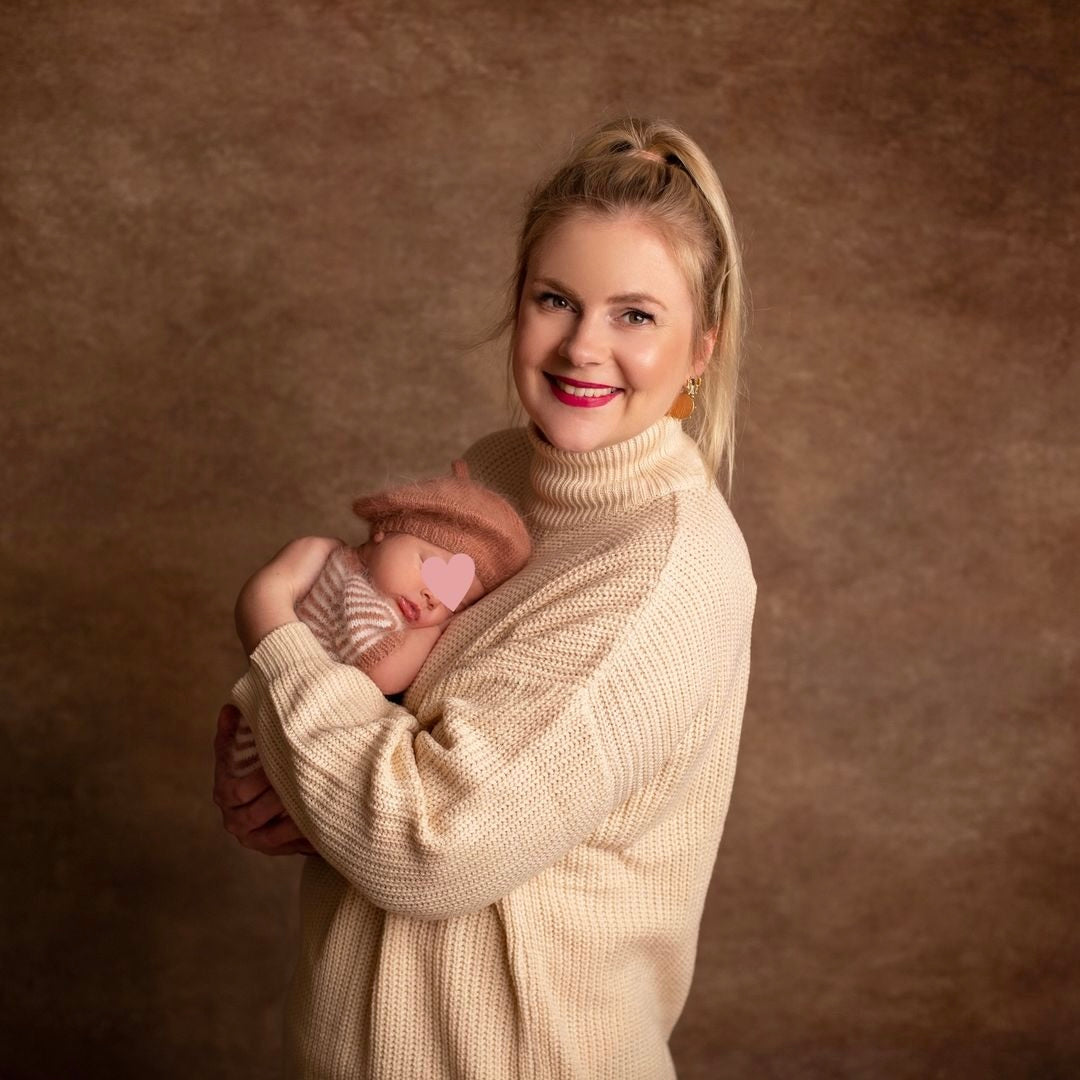 Eine Frau in beigem Pulli hält ein Baby in einem rosa Strickoutfit. Warmes, leicht verschwommenes Hintergrundbild.