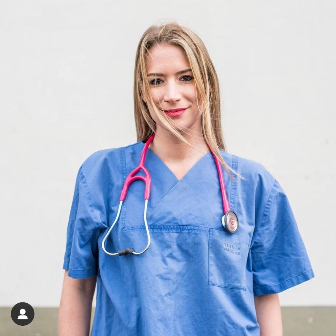 Junge Frau in blauer Arztuniform mit Stethoskop, lächelnd und mit langen, offenen Haaren. Hintergrund hell.