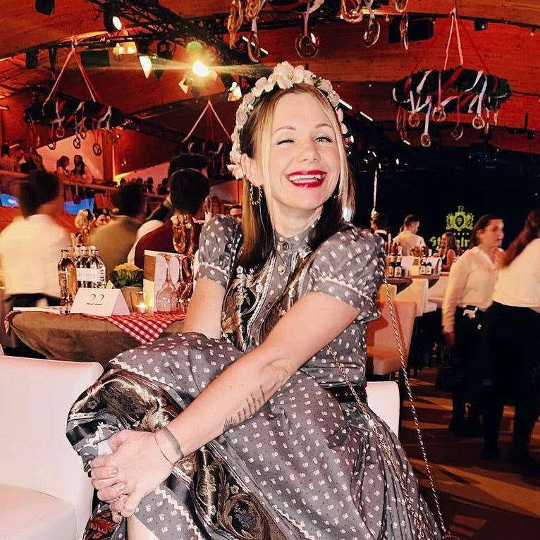 Eine lächelnde Frau in einem gepunkteten Kleid mit Blumenkranz sitzt auf einem eleganten Event, umgeben von weiteren Gästen.