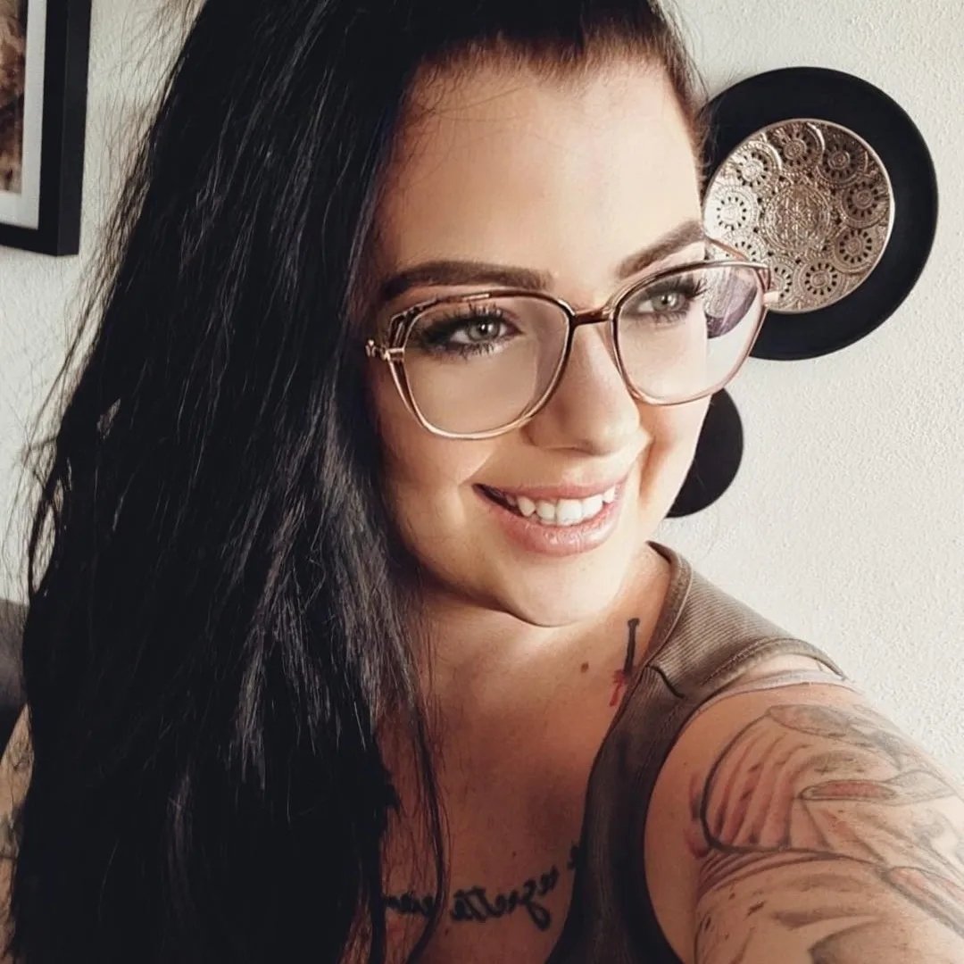 Lächelnde Frau mit langen, dunklen Haaren und Brille, tätowierte Arme, entspannte Atmosphäre.