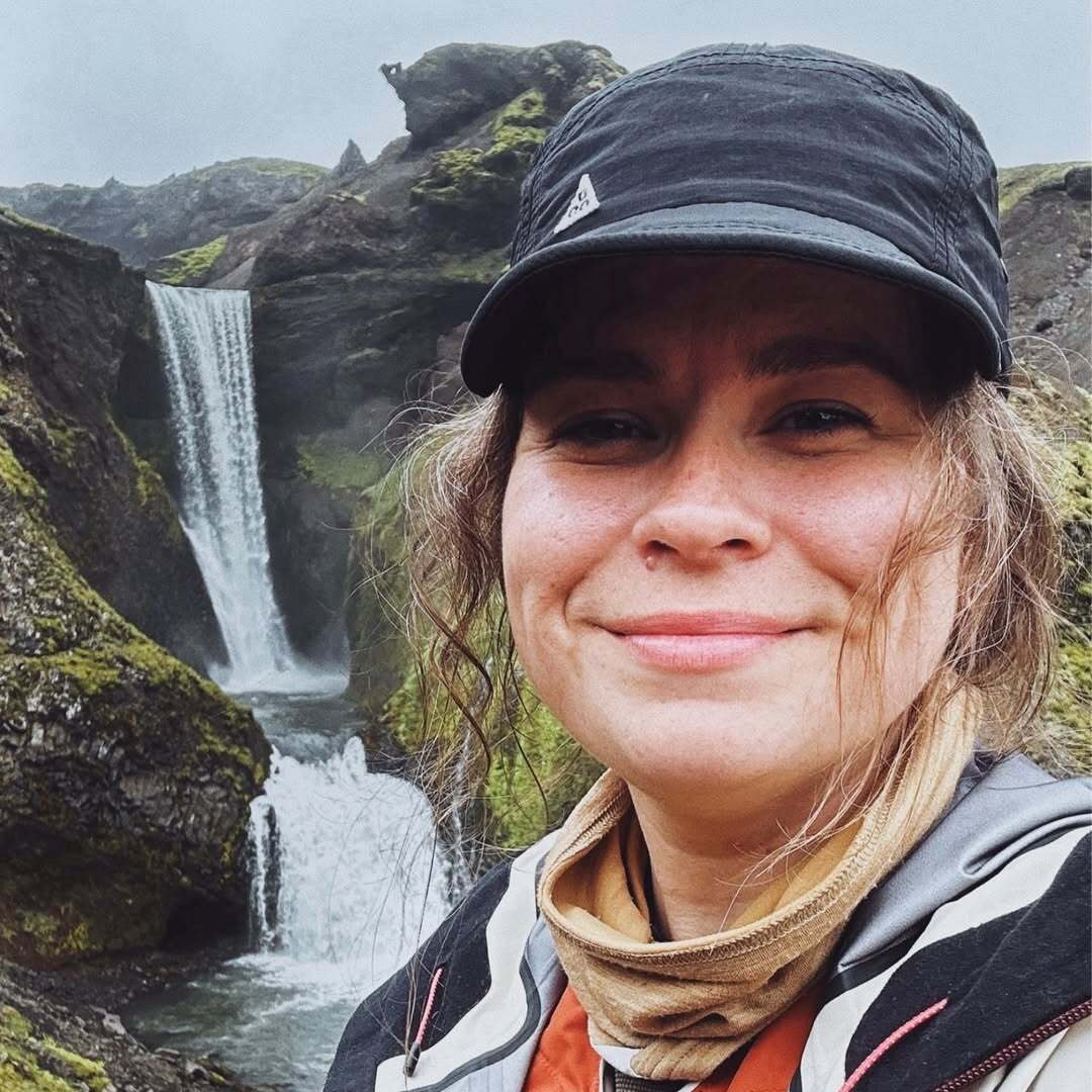 Eine Frau in sportlicher Kleidung und Cap steht lächelnd vor einer beeindruckenden Wasserfall-Landschaft in der Natur.