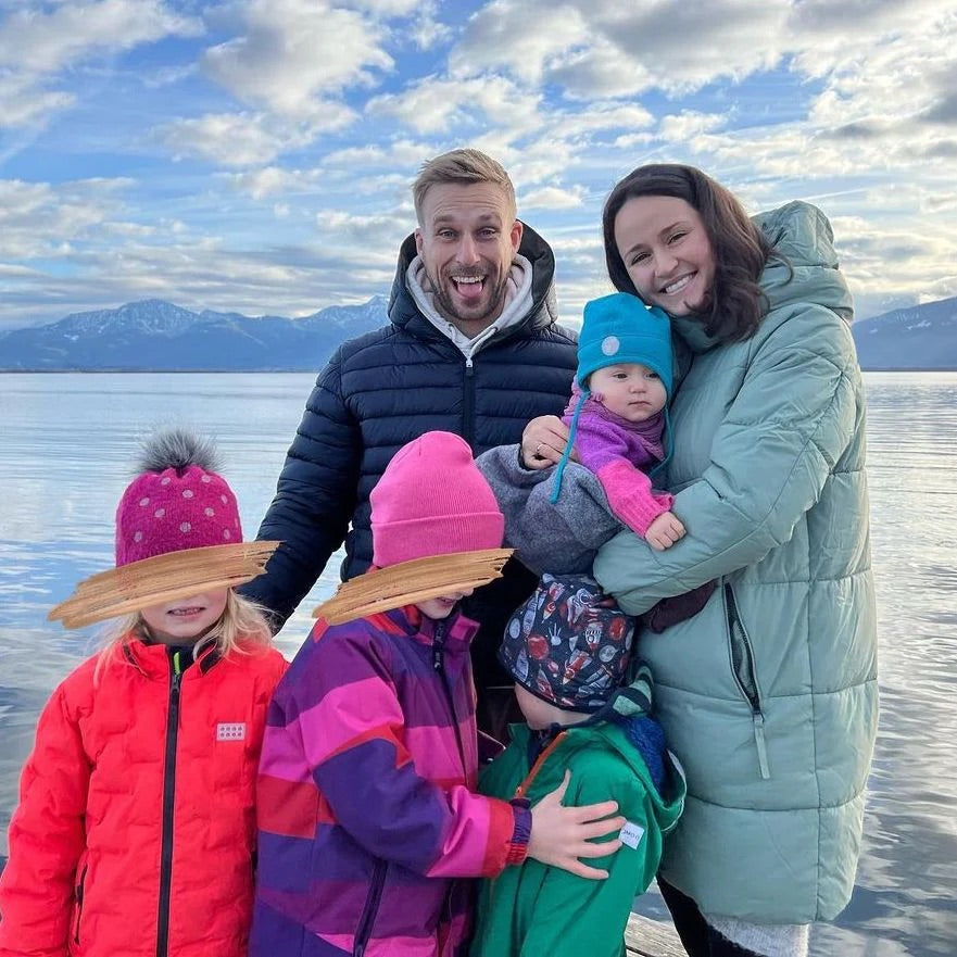 Eine Familie steht am Wasser. Die Eltern umarmen die Kinder, die in bunten Jacken und Mützen gekleidet sind. Blick auf Berge