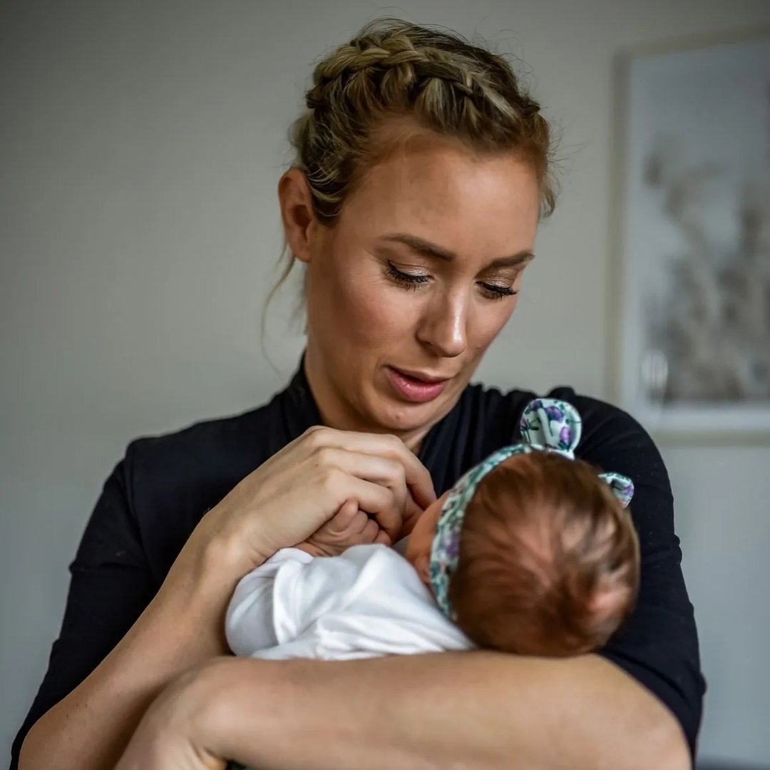 Eine Mutter hält ihr Neugeborenes im Arm und schaut liebevoll auf es. Sie trägt ein schwarzes Oberteil und hat einen