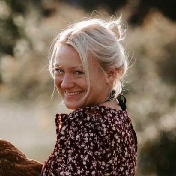 Eine lächelnde Frau mit blonden Haaren, die in einem blumigen Kleid in einer natürlichen Umgebung steht.