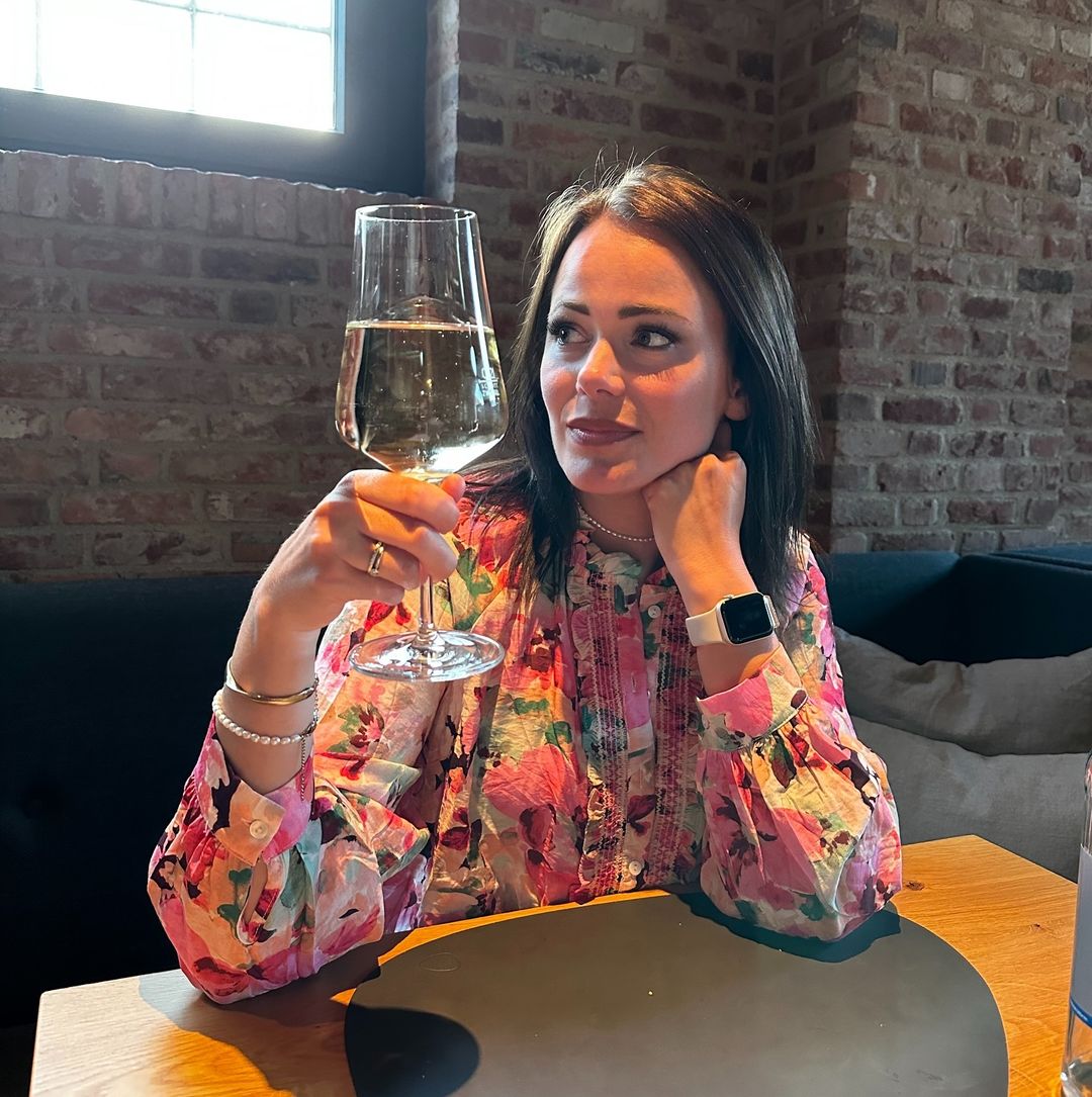 Eine Frau mit blühender Bluse hält ein Glas Weißwein. Sie sitzt in einem gemütlichen, brick-walled Café.