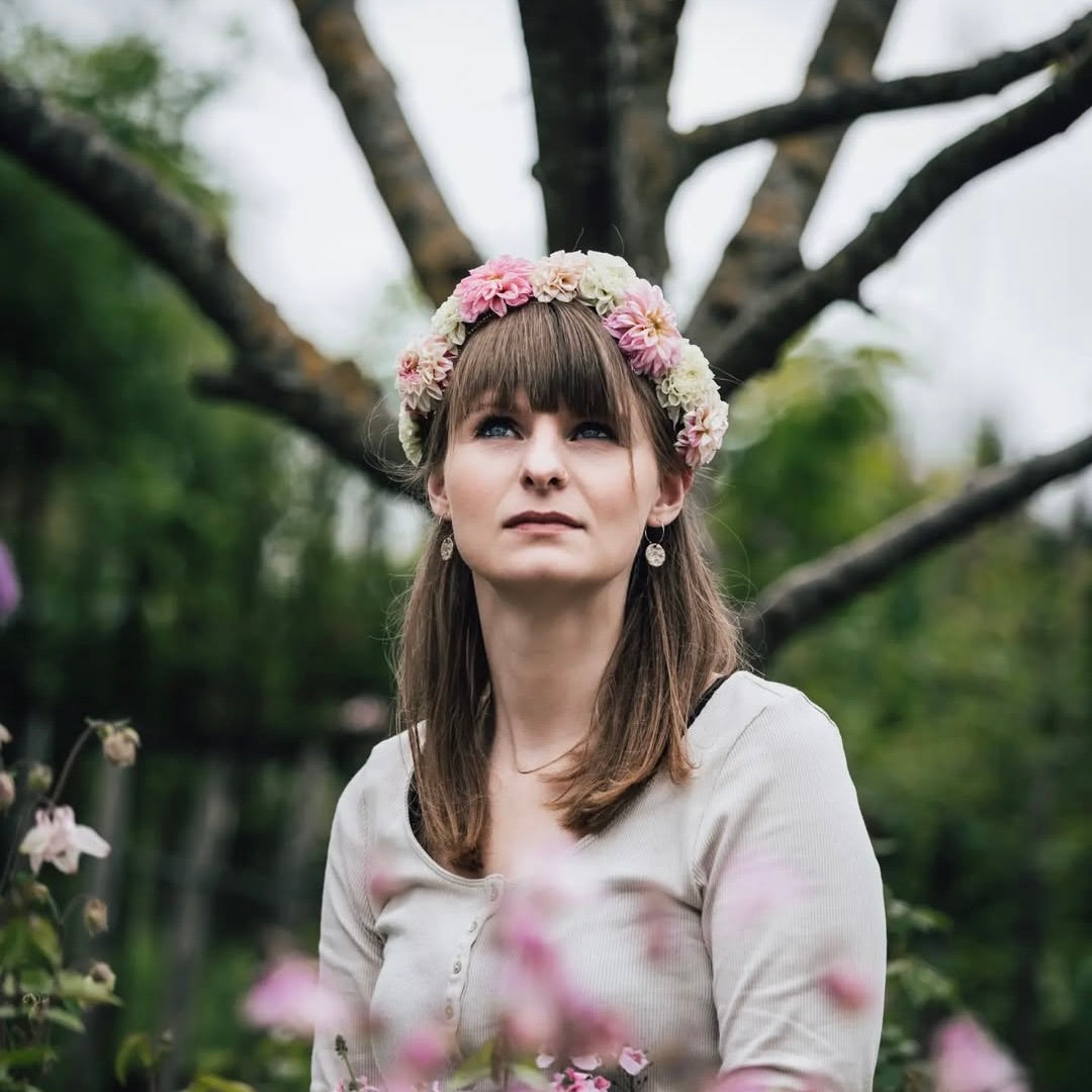 Frau mit Blumenkranz und schlichter Bluse sitzt in einem blühenden Garten, umgeben von Pflanzen.
