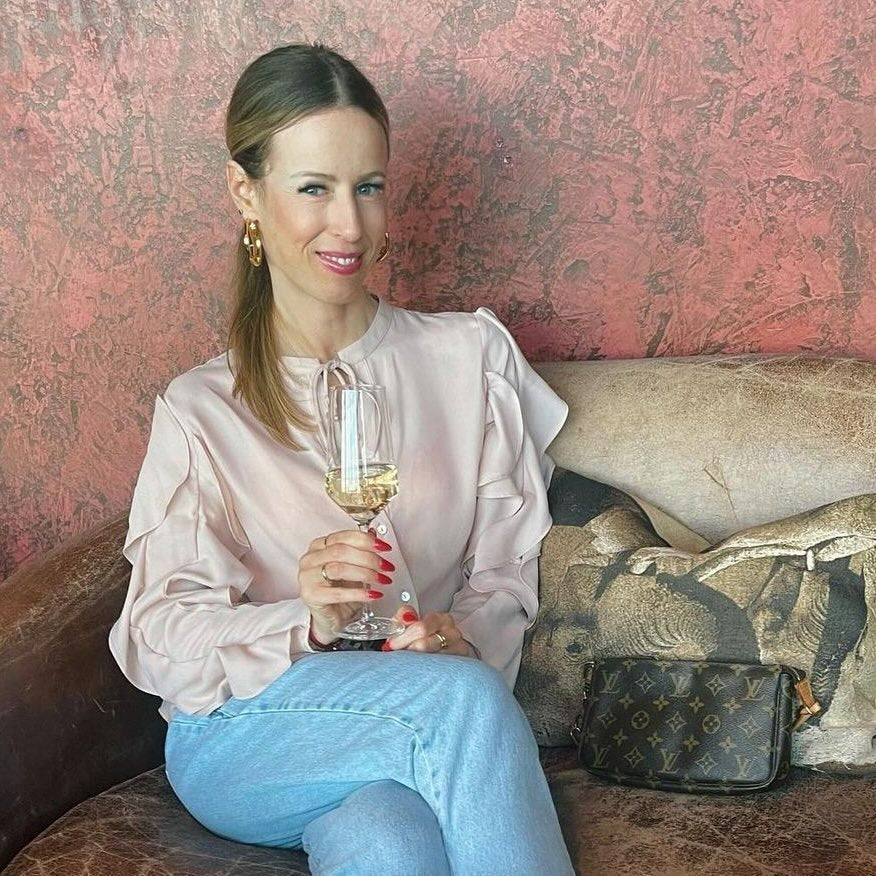 Eine Frau mit blonden Haaren in einem roséfarbenen Oberteil sitzt auf einer Couch und hält ein Glas Sekt. Sie trägt goldene