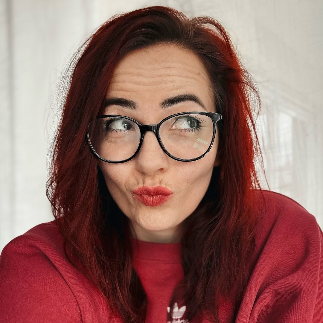 Ein Porträt einer Frau mit rotem Haar und Brille, sie trägt ein rotes Sweatshirt und zeigt einen nachdenklichen